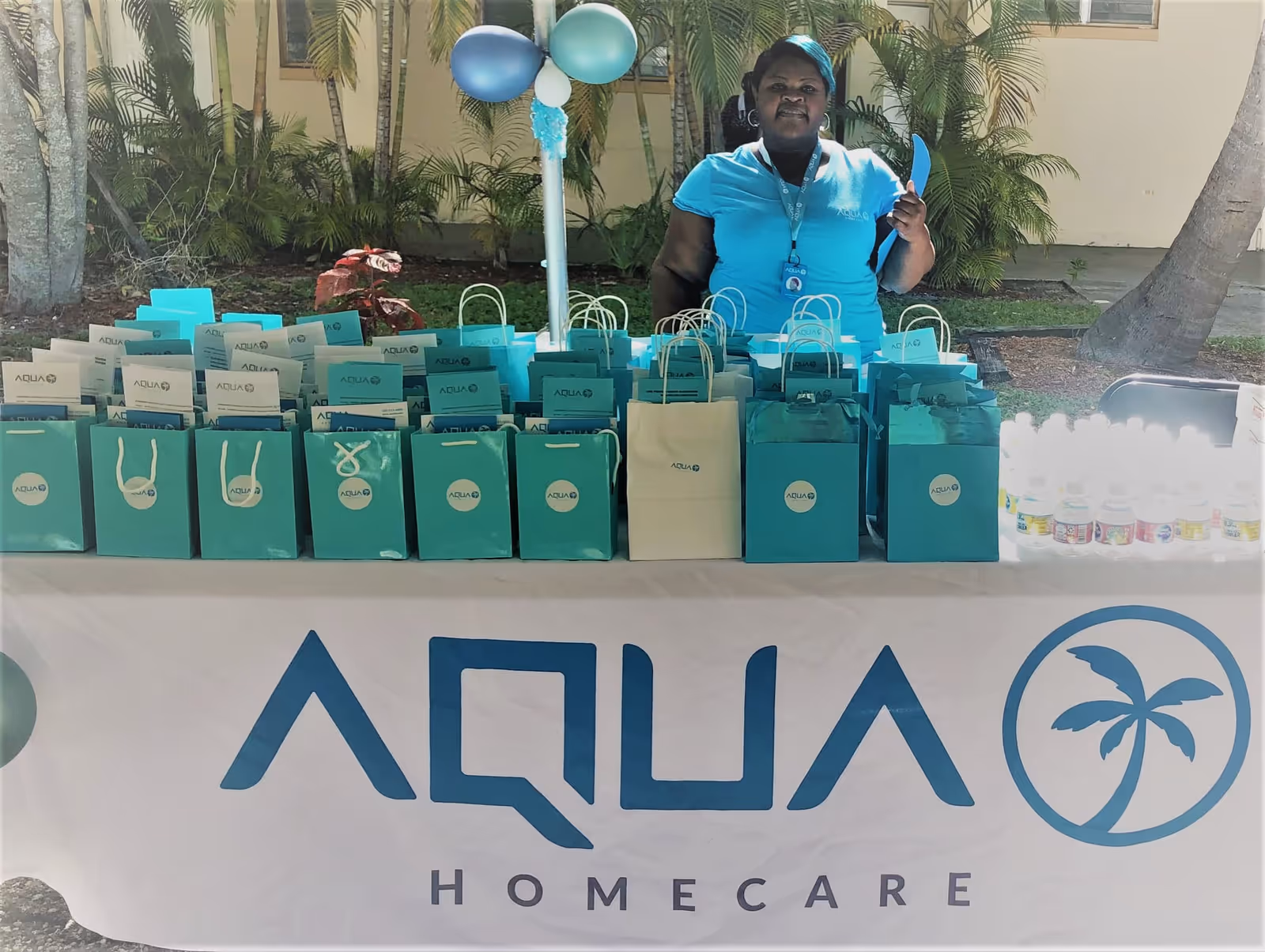 Staff in blue uniform stands behind a table with aqua gift bags outdoors, palm trees visible.