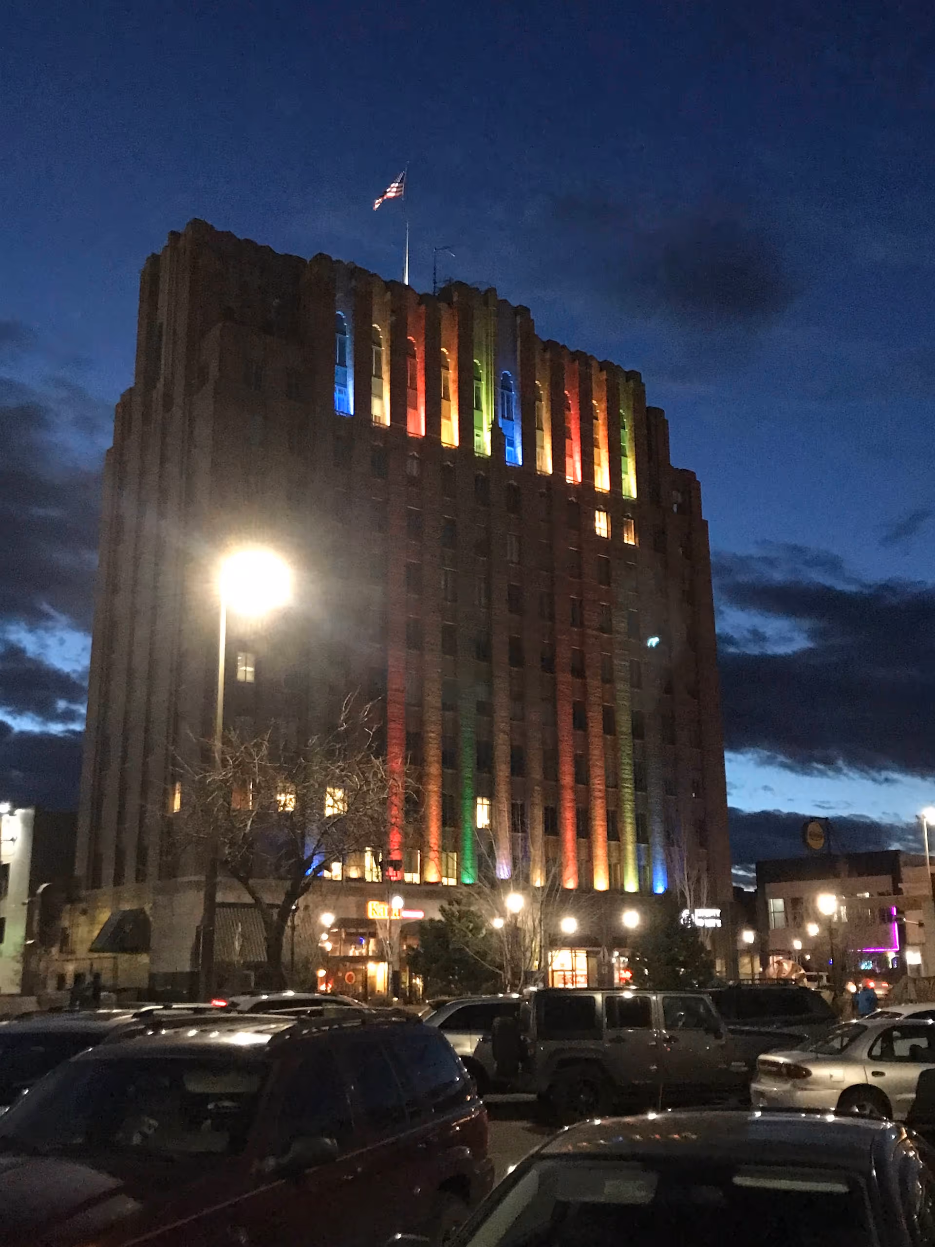 Night view of a tall building with rainbow-lit façade and surrounding parked cars.