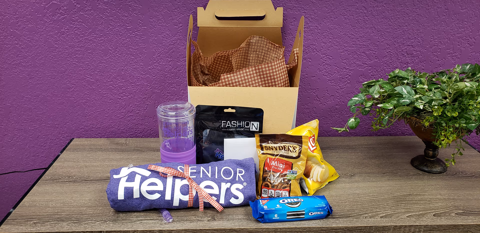 A table with snacks, a gift box, tumbler, and a potted plant against a purple wall.