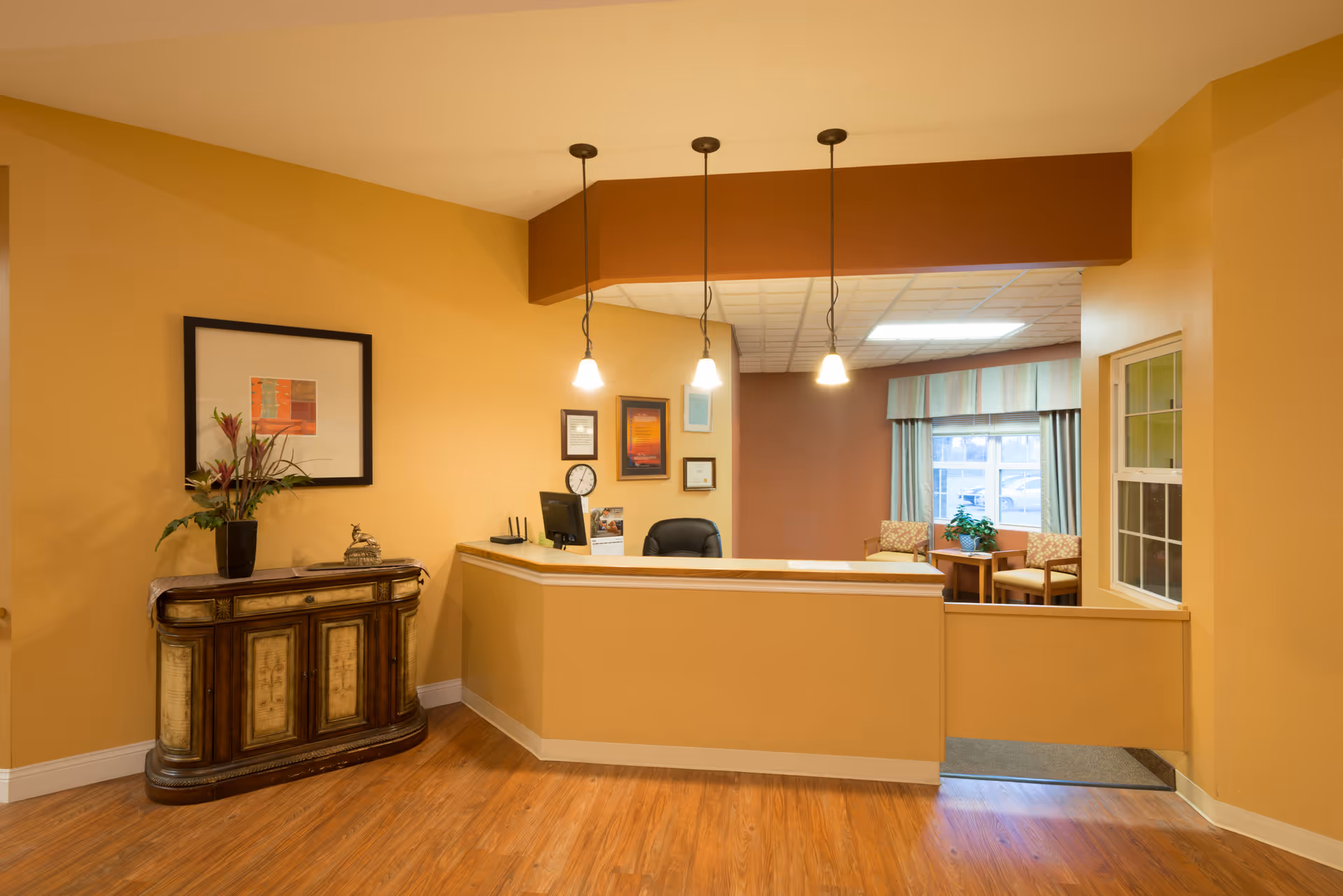 Reception area with a wooden floor and a beige and brown color scheme. There is a reception desk with three hanging pendant lights above it, a computer monitor, and a black office chair behind the desk. To the left, there is a decorative wooden cabinet with a vase of flowers and a framed artwork on the wall above it. In the background, there is a seating area with two chairs, a small table, and a window with curtains.