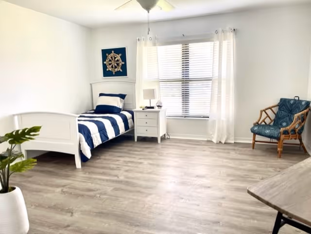 A bright and clean bedroom with a single white bed featuring navy blue and white striped bedding. Next to the bed is a white nightstand with a lamp and a small decorative item. A large window with white curtains allows natural light to fill the room. There is a blue cushioned chair with a wooden frame in the corner and a green potted plant in a white pot on the left side. The floor is light-colored wood, and the walls are painted white with a nautical-themed decoration above the bed.