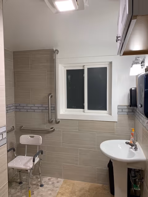Accessible bathroom with grab bars, a shower chair, sink, and window.