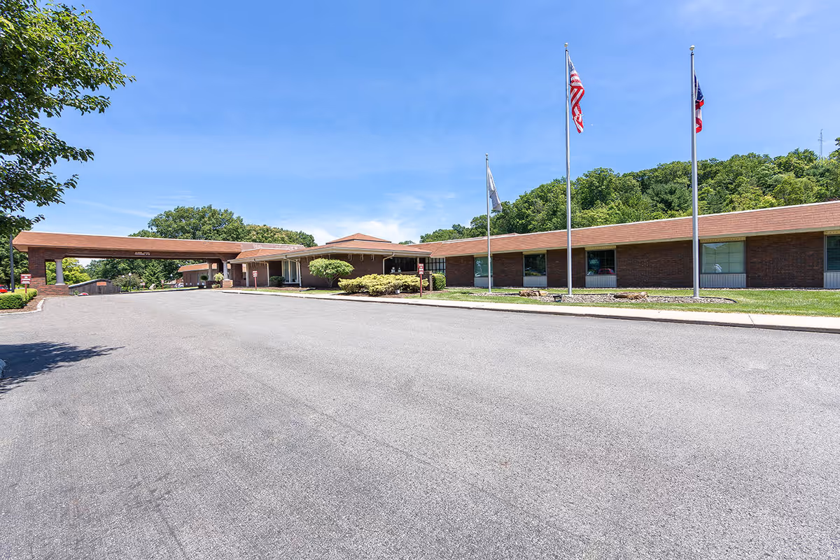 Front exterior of a single-story brick senior living facility with a covered drive-through entrance, a paved driveway, and three flagpoles.