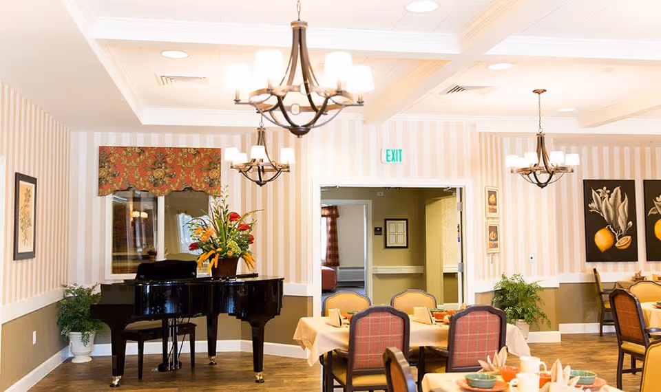 A dining room in a senior living facility with tables set for a meal, upholstered chairs, a black grand piano with a floral arrangement on top, striped wallpaper, framed artwork, and three chandeliers hanging from the ceiling.
