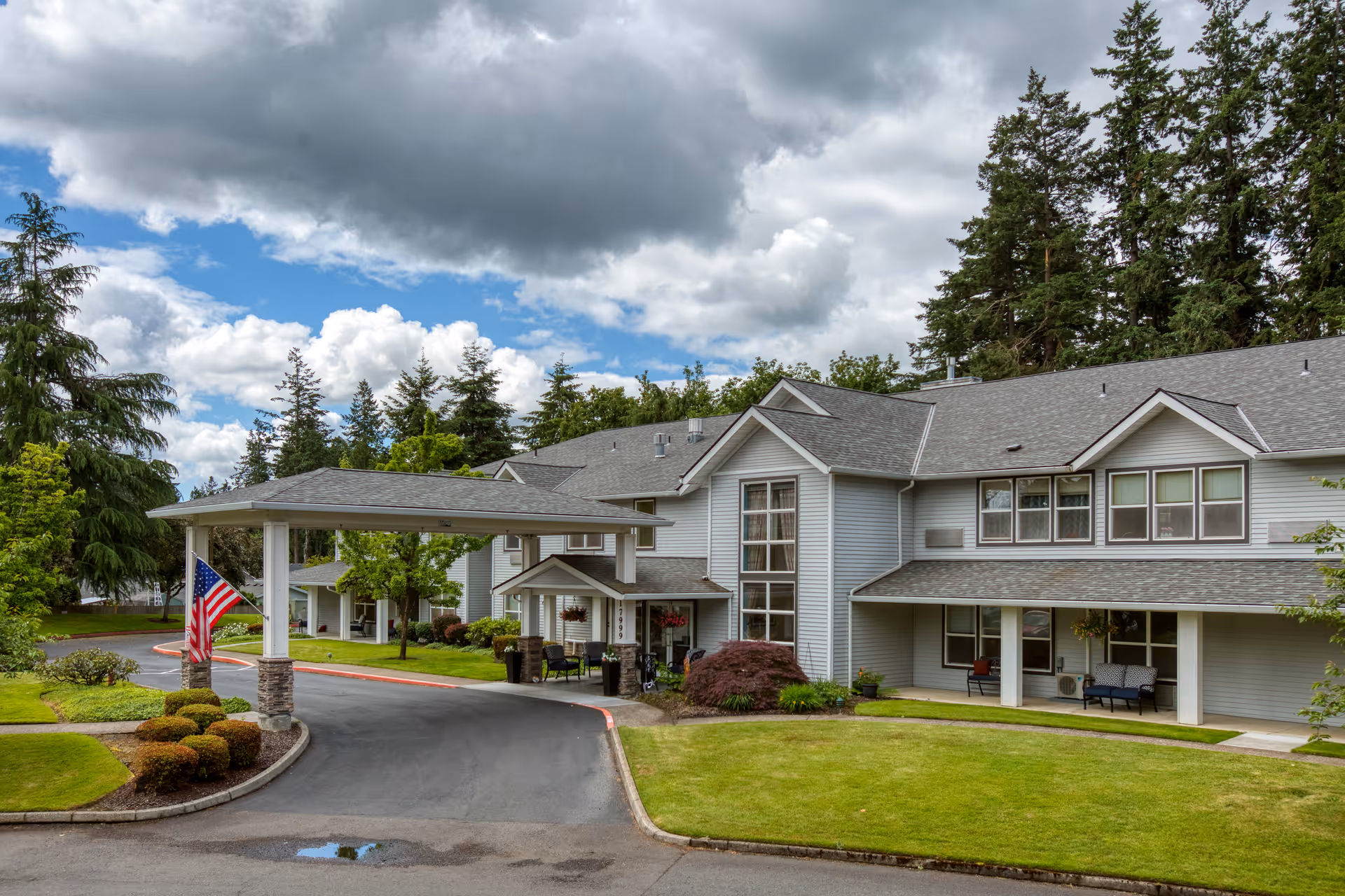 Exterior view of Homewood Assisted Living facility showing a two-story building with gray siding and a covered entrance. The driveway curves around landscaped bushes and green lawns, with tall trees in the background under a partly cloudy sky.