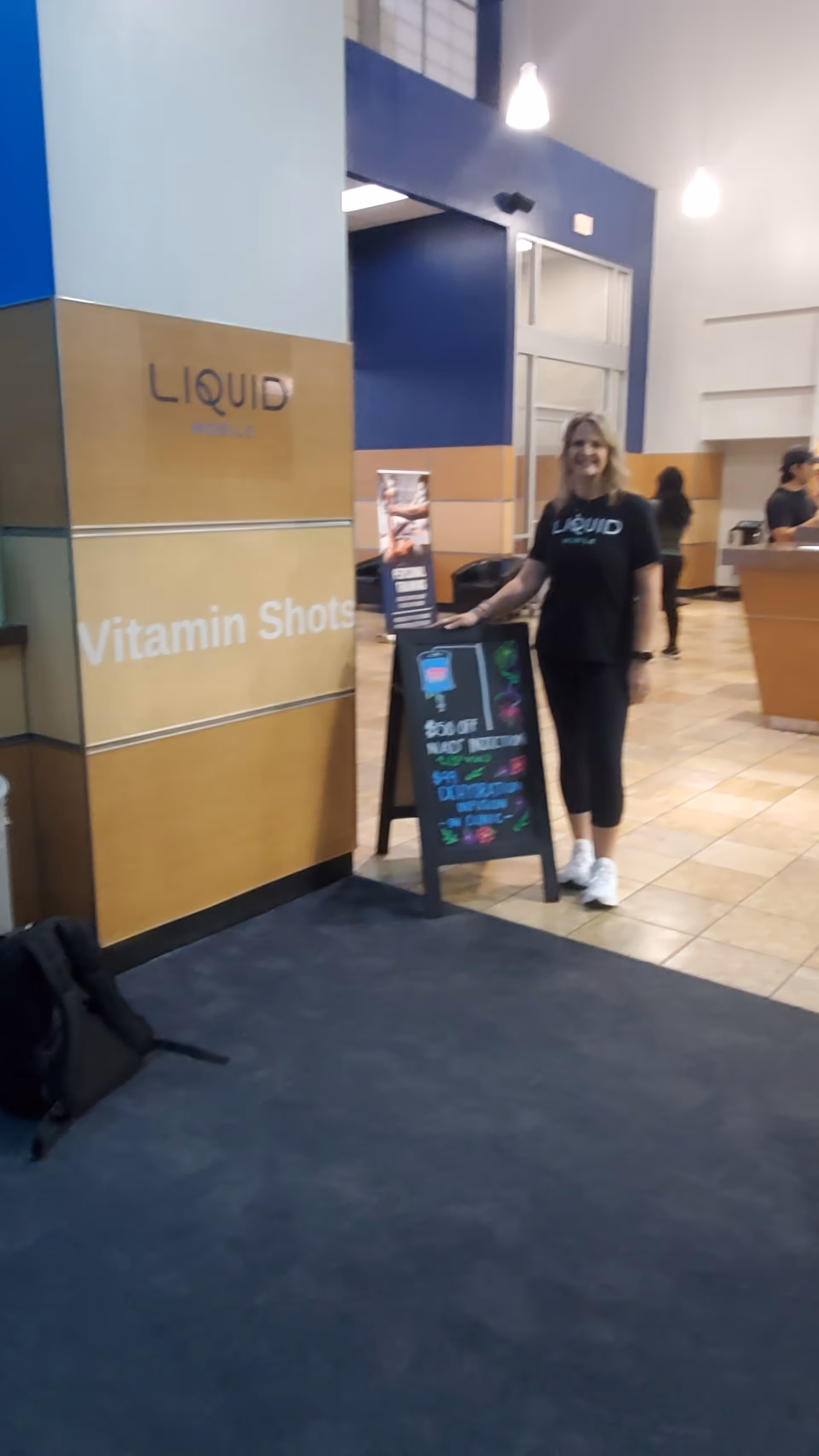 Staff member in Liquid Mobile IV shirt stands beside a chalkboard sign indoors.