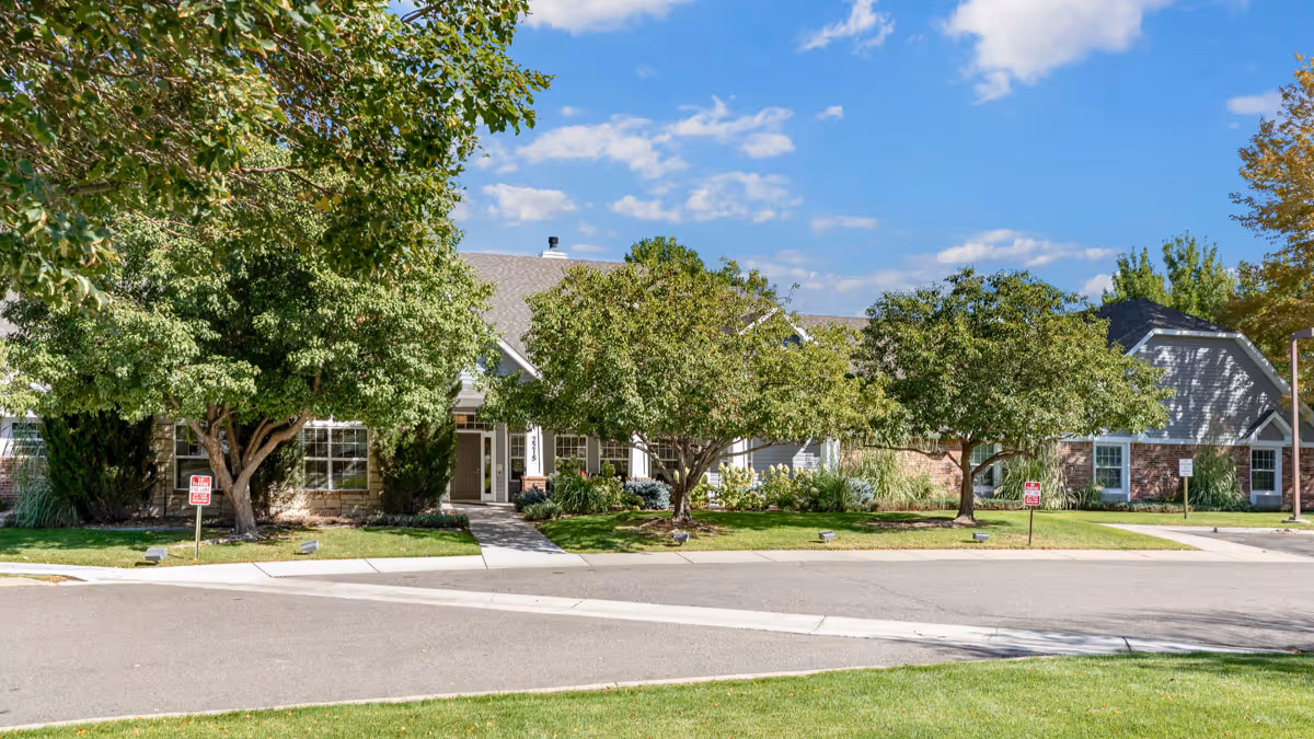 Single-story senior living facility facade with mature trees and a sidewalk along the street.