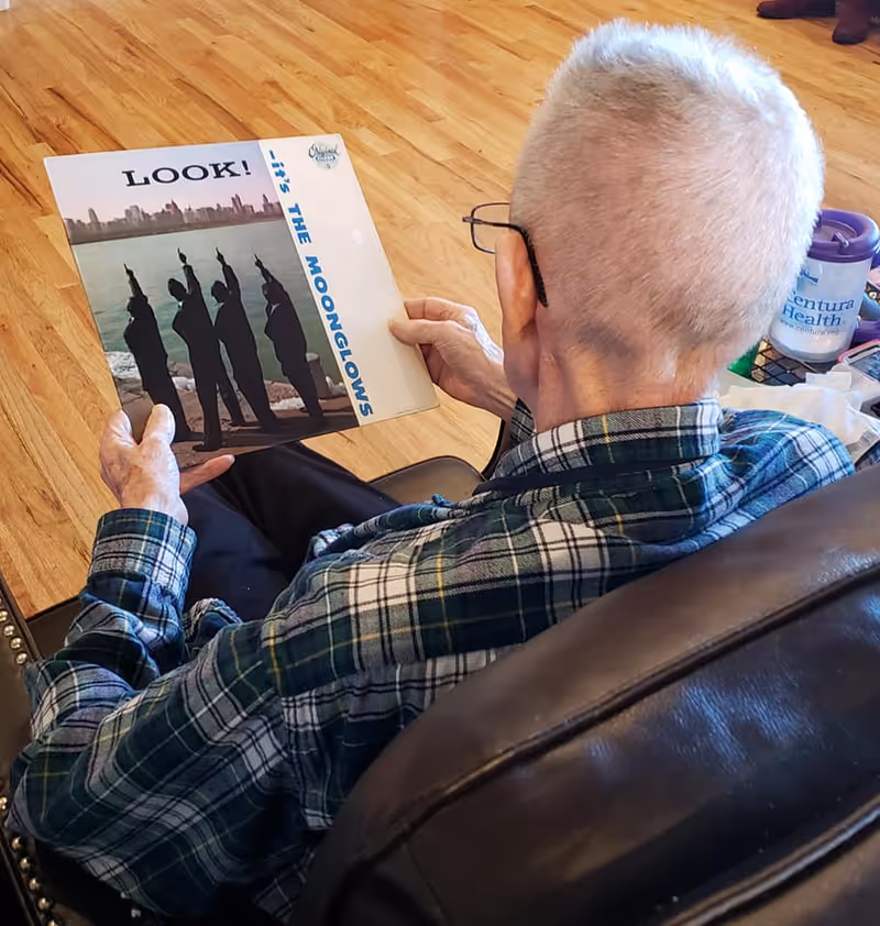 An elderly person with short white hair and glasses is sitting in a dark leather chair, holding and looking at a vinyl record album titled 'LOOK! It's The Moonglows'. The person is wearing a green and white plaid shirt. The floor is wooden, and there is a table nearby with a Ventura Health cup and some other items.