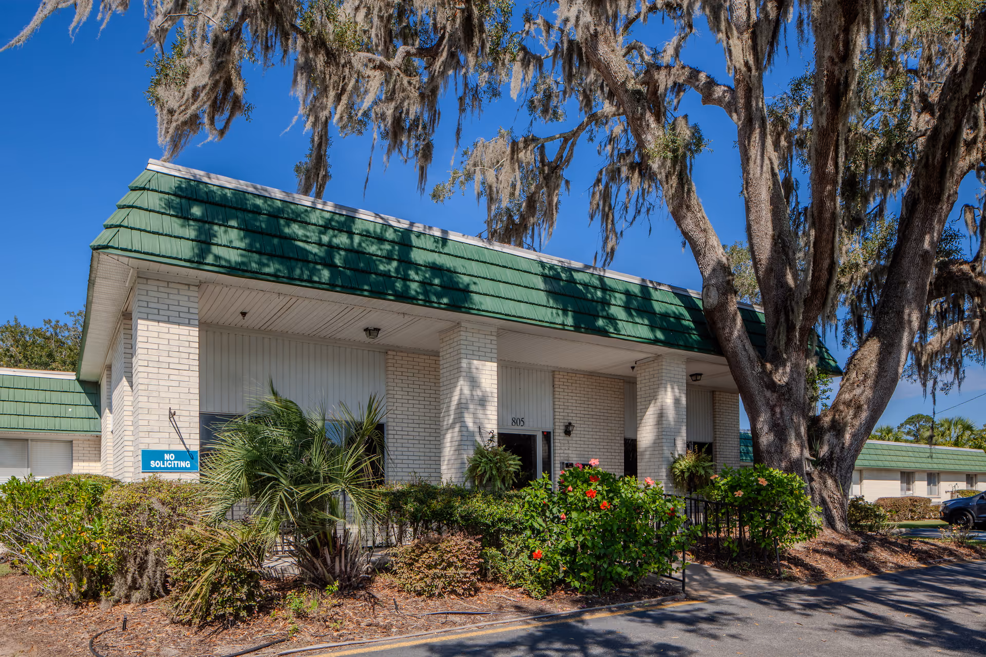 Exterior view of a white brick building with a green roof and large trees, landscaped front.