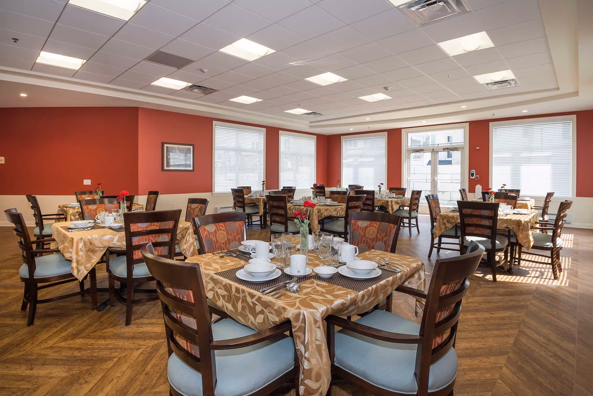 A bright dining room with multiple tables covered in beige patterned tablecloths, each set with white dishes, cups, glasses, and silverware. The chairs have wooden frames with patterned backs and light blue cushions. The room has large windows with white blinds, red walls, and a wood-patterned floor.