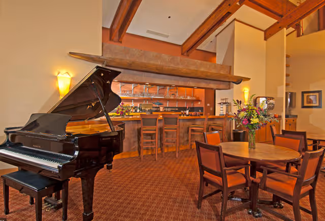 Elegant senior living lounge with grand piano, bar, and round dining table.