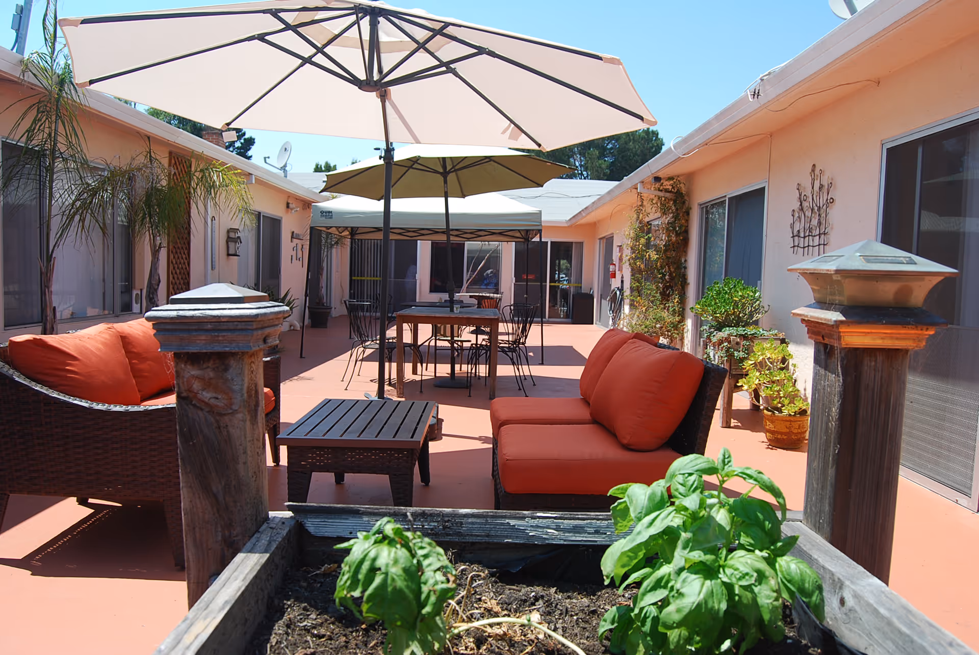 Sunny outdoor courtyard with red-cushioned lounge seating, tables and umbrellas, potted plants, and surrounding single-story rooms.