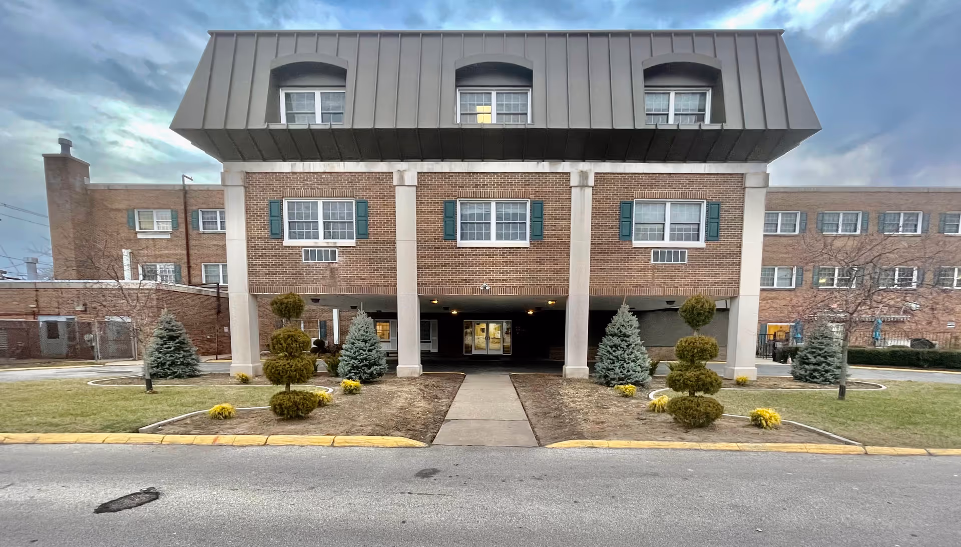 Front exterior view of a multi-story brick building with a covered entrance supported by columns, landscaped with trimmed bushes and small trees, under a cloudy sky.