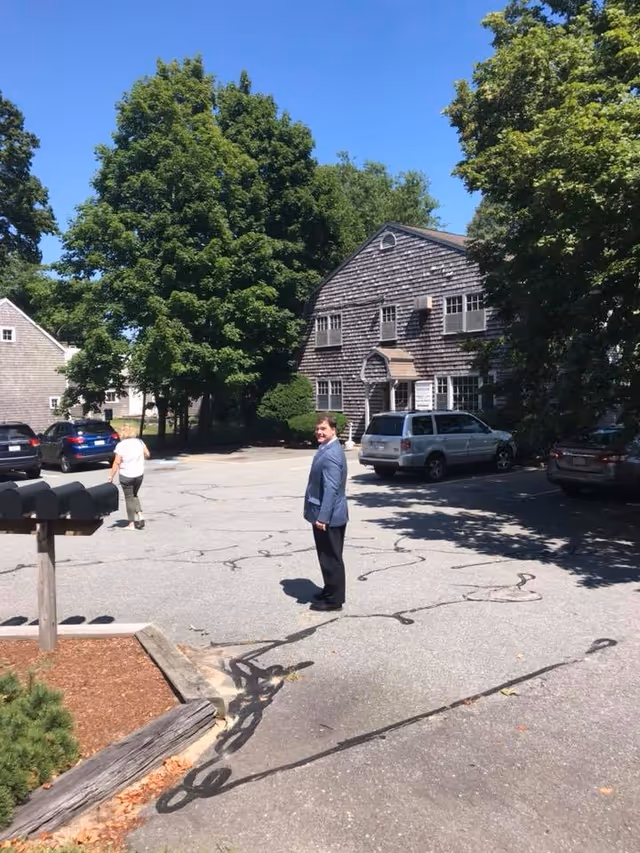 Senior man in blazer stands in parking lot outside a wooden care facility.