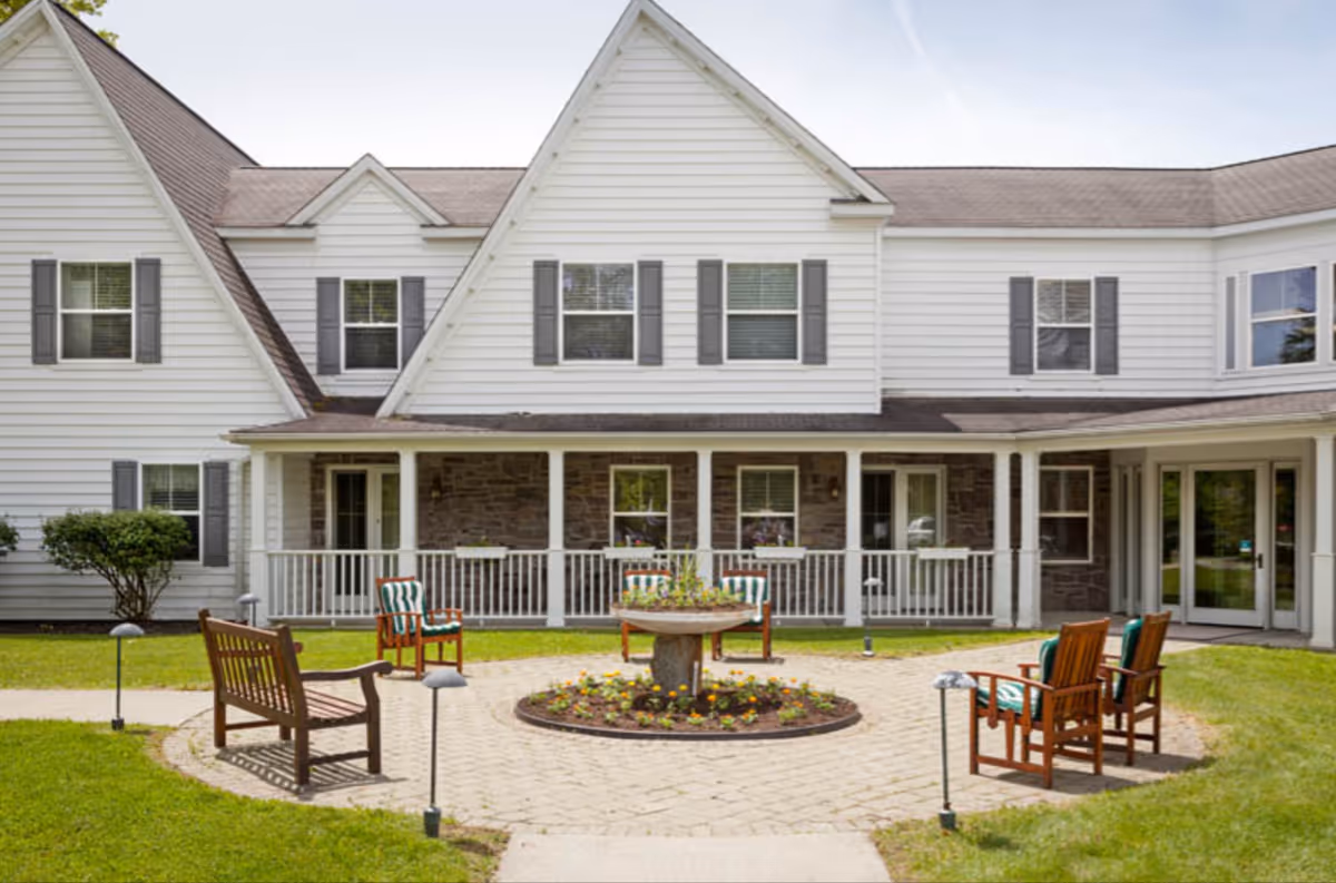 Outdoor patio area at The Inn At Millbrook featuring wooden chairs with cushions arranged around a circular flower bed on a paved surface, in front of a white building with gray shutters and a porch.