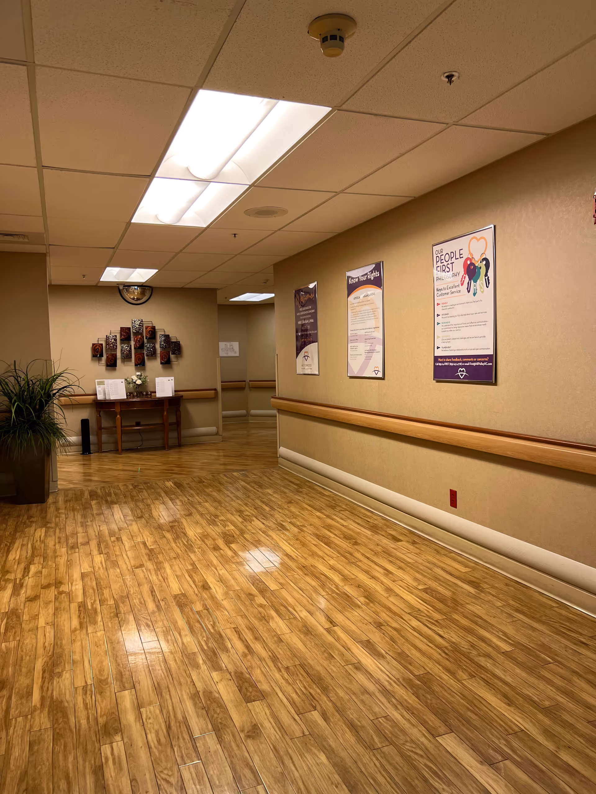 Long indoor hallway with wood-like flooring, beige walls, and wall posters.