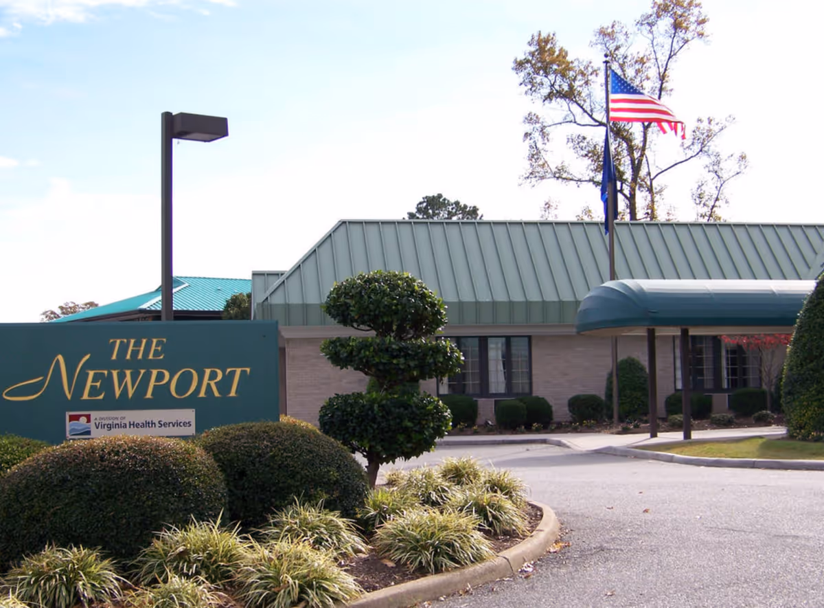 Exterior view of The Newport Nursing and Rehabilitation Center building with a green metal roof, an American flag flying on a flagpole, manicured bushes, and a sign that reads 'The Newport, a division of Virginia Health Services'.