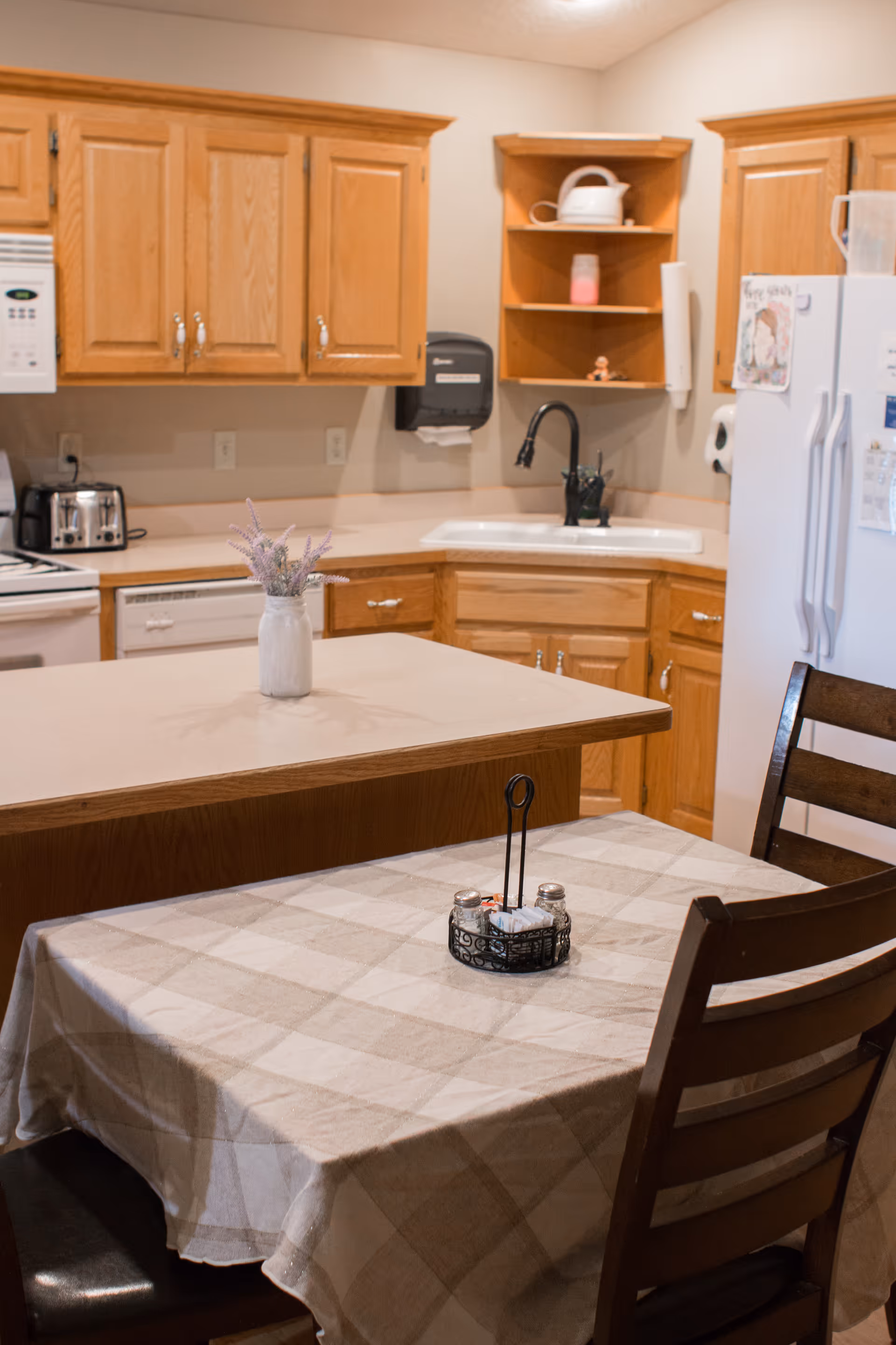 A kitchen area with wooden cabinets, a white countertop island with a vase of flowers, a sink with a black faucet, a white refrigerator, and a small dining table covered with a checkered tablecloth and two wooden chairs. The table has a small condiment holder with salt, pepper, and napkins.