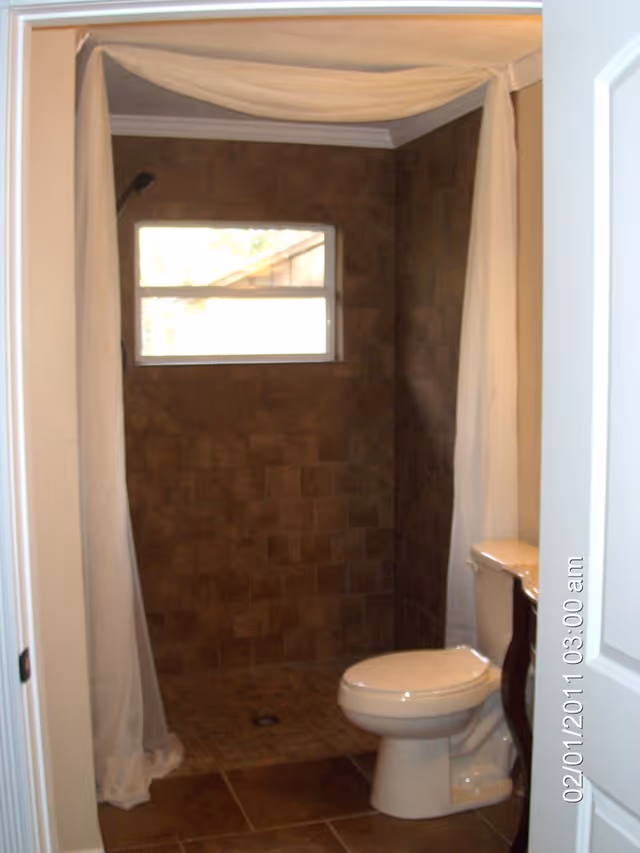 Bathroom with a tiled walk-in shower area featuring a small window, a white toilet, and a white shower curtain partially drawn to the side.