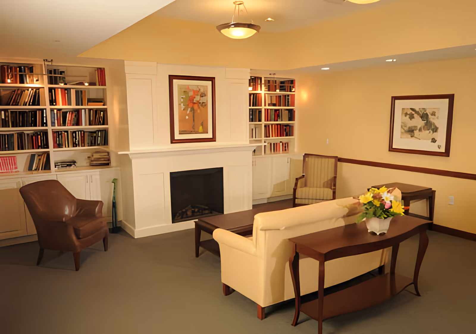 A cozy living room area with a beige sofa, a wooden coffee table, a side table with a vase of flowers, a brown armchair, and a striped armchair. The room features built-in white bookshelves filled with books on either side of a white fireplace with a framed artwork above it. The walls are painted a warm beige color and there is another framed artwork on the right wall.