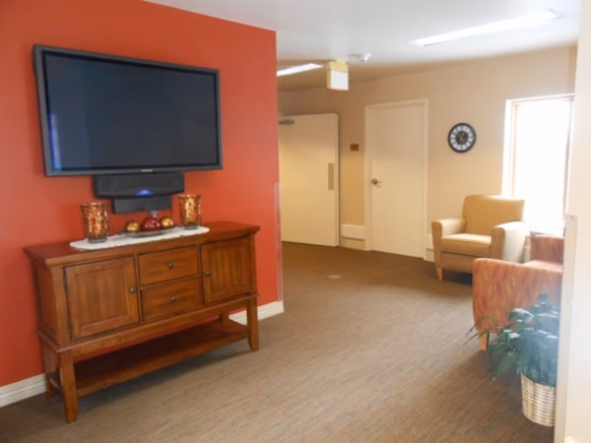 A cozy interior room with a wall-mounted flat screen TV above a wooden cabinet decorated with candles and ornamental balls. The room has a red accent wall behind the TV and beige walls elsewhere. There are two armchairs near a window, a wall clock, and a potted plant in a basket on the floor.