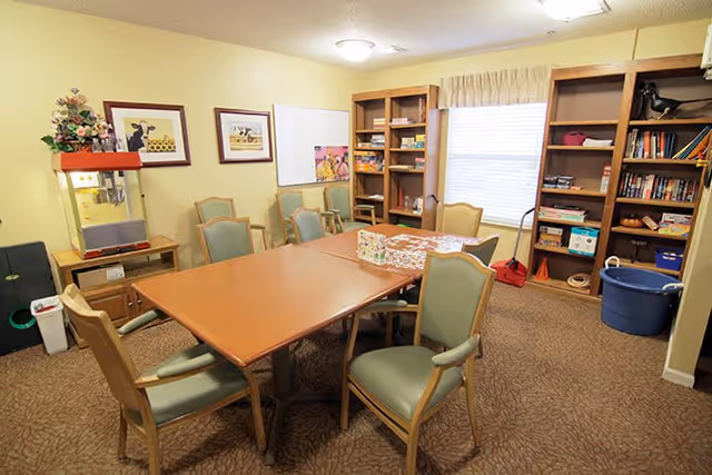 A cozy activity room with a rectangular wooden table surrounded by six green cushioned chairs. The room has beige walls and carpeted floor. On the left side, there is a popcorn machine on a small wooden cabinet, and two framed pictures hang on the wall above it. The back wall features a large window with beige curtains and two wooden bookshelves filled with games, books, and various items. A puzzle is partially completed on the table.