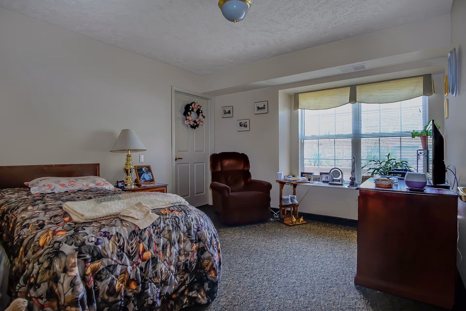 Bedroom in Grandhaven Living Center with floral bedspread, lamp, chair, and large window.
