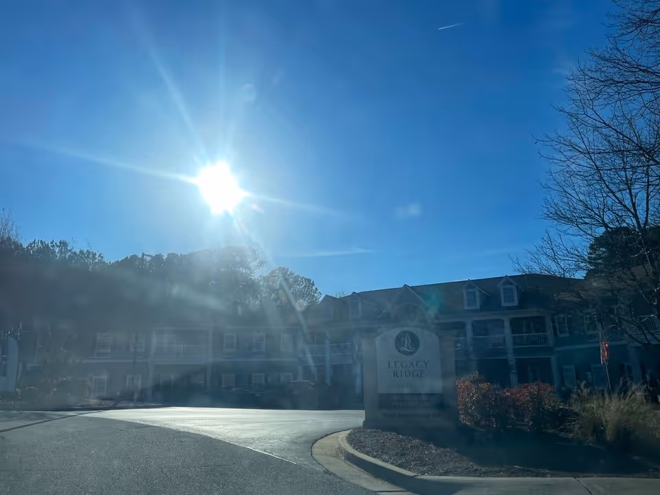 Sunlit front exterior of a multi-story senior living building with a driveway and a sign reading 'Legacy Ridge'.