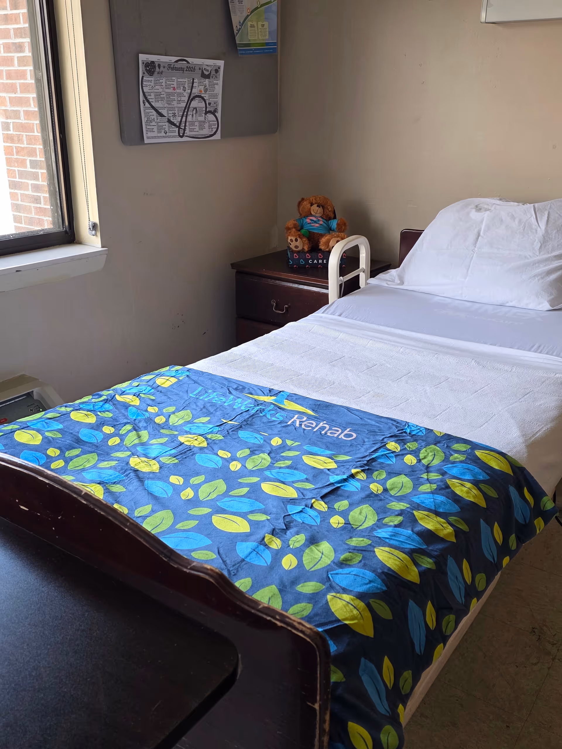 A hospital-style patient room with a bed, nightstand, teddy bear, and window.