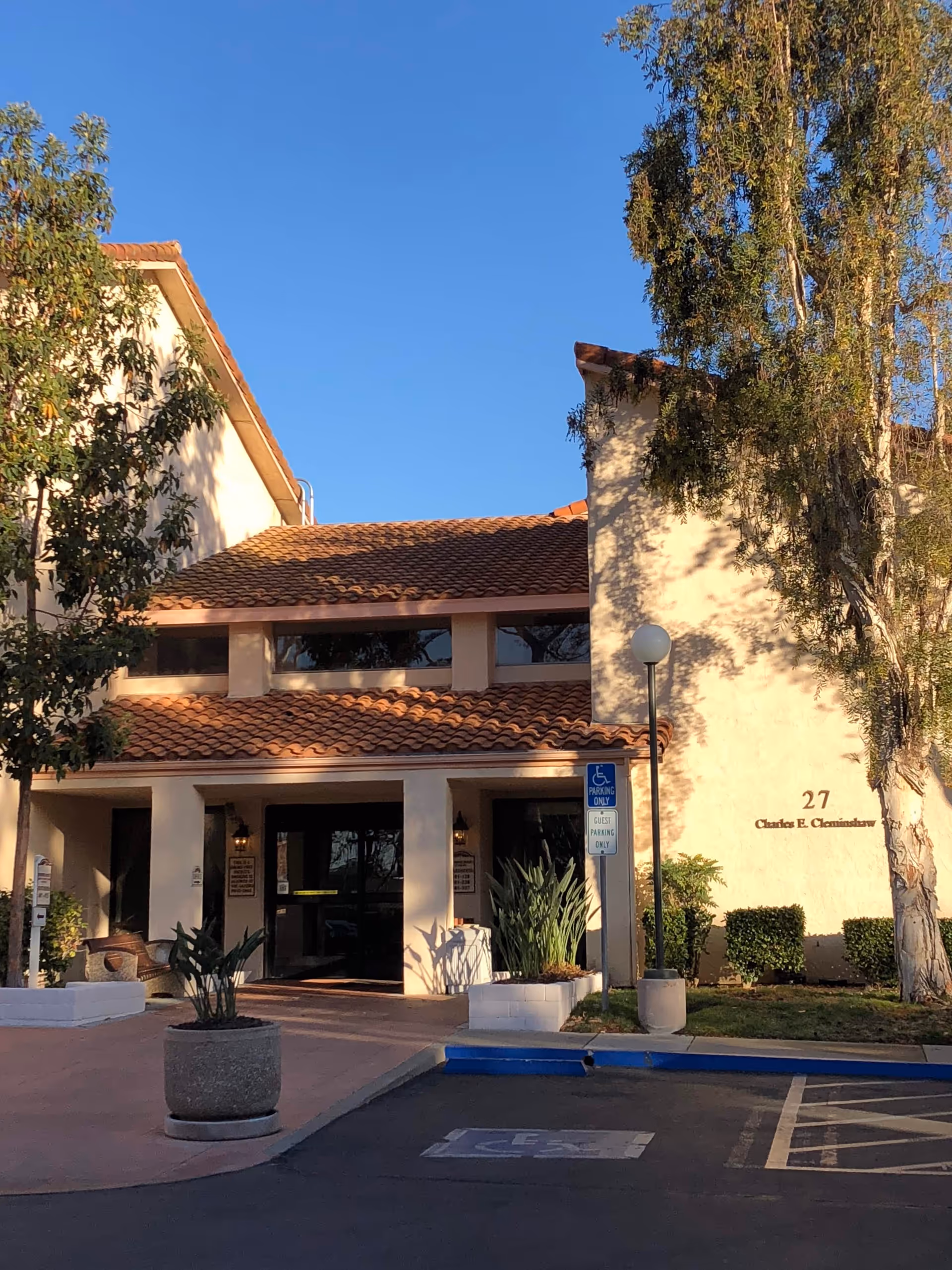 Front exterior of Woodbridge Manor with beige stucco, red tiled roof, palm trees, and parking area.