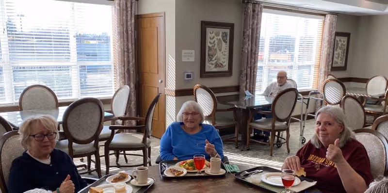 Three elderly women sitting at a dining table in a well-lit room with large windows and beige curtains. They have meal trays in front of them with food and drinks. One woman is giving a thumbs-up gesture. In the background, an elderly man is seated at another table near the window.