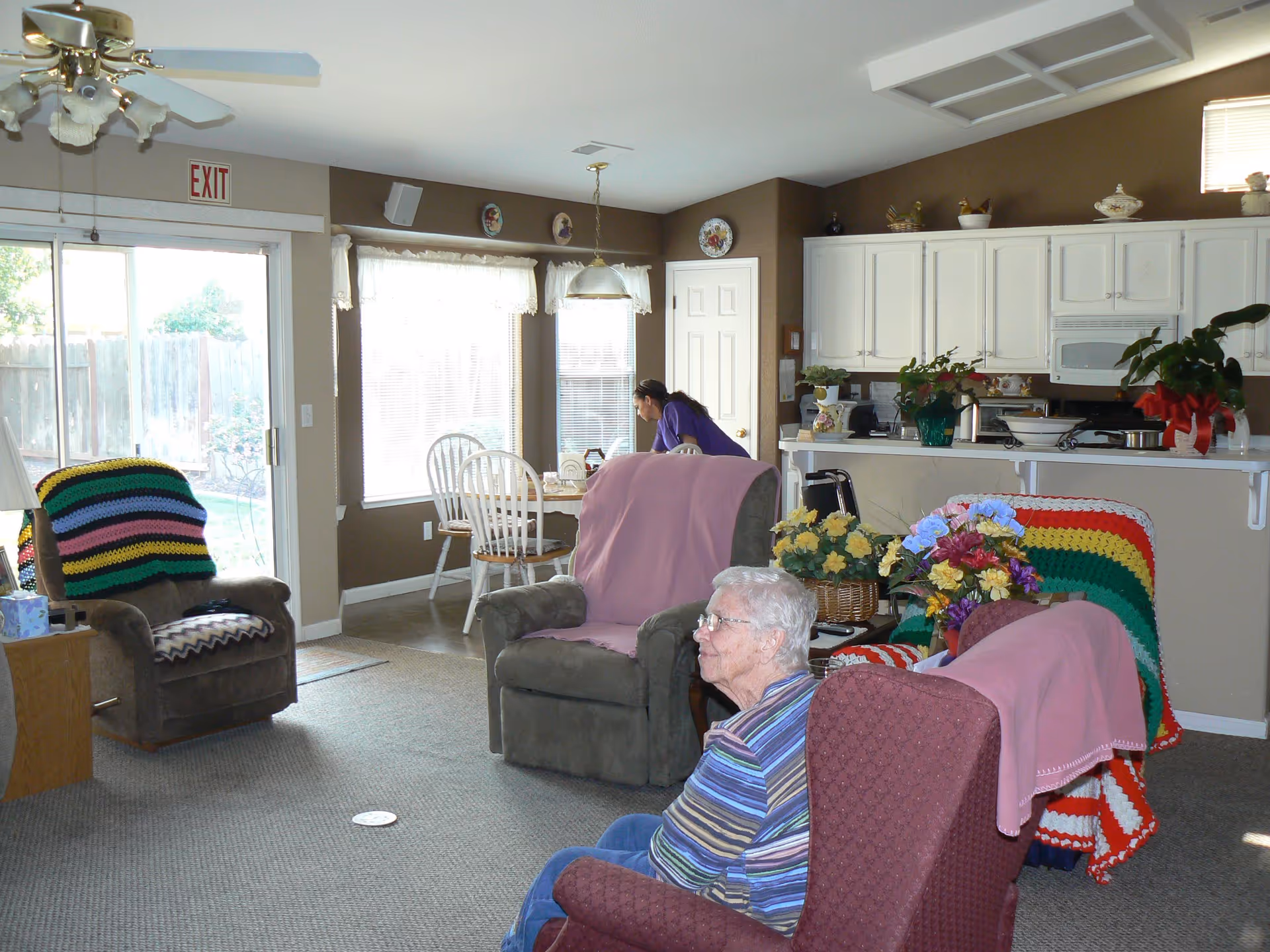 A cozy living room area in a senior care facility with several armchairs covered with colorful blankets. An elderly woman is sitting in one of the chairs. In the background, a caregiver is seen near a dining table by large windows with sheer curtains. The kitchen area with white cabinets and various plants and decorations is visible behind the dining area.