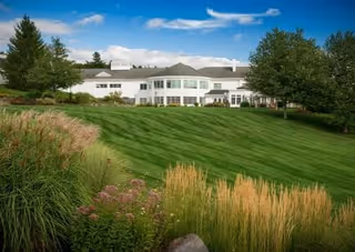 A large white building with multiple windows situated on a grassy hill surrounded by trees and ornamental grasses under a blue sky with some clouds.