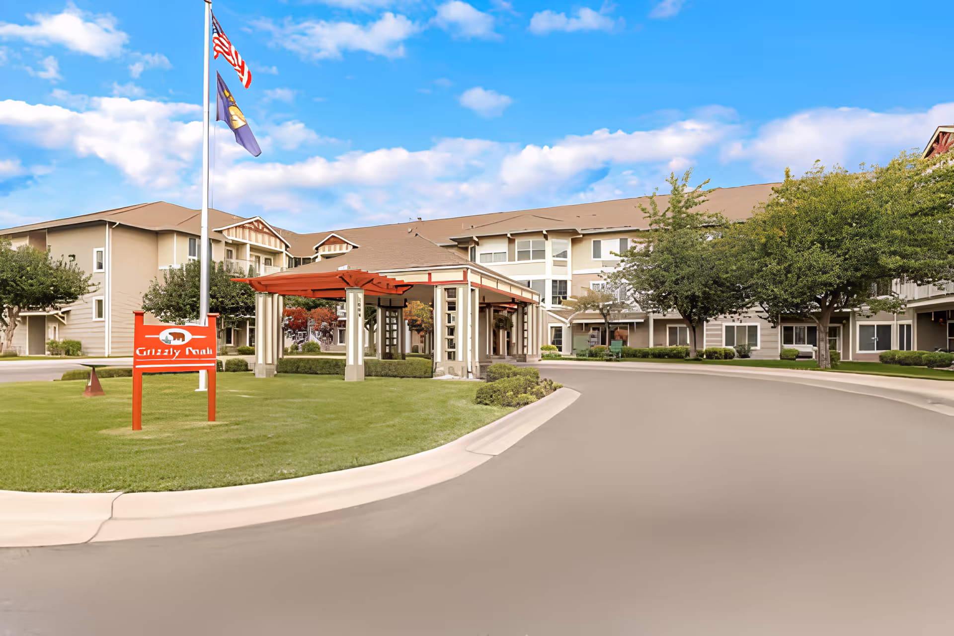 Exterior view of a senior living facility named Grizzly Peak, featuring a large building with multiple windows, a covered entrance with columns, a well-maintained lawn, trees, and two flagpoles displaying the American flag and another flag.