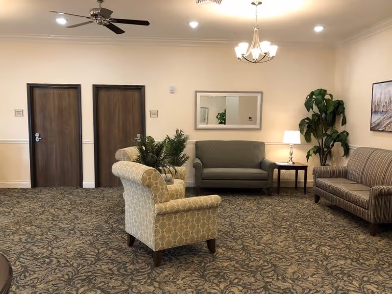 Carpeted common lounge with armchairs, sofas, side tables and plants arranged under ceiling lights.