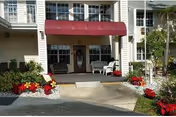 Front entrance of Bayou Gardens Assisted Living with maroon awning, chairs, and colorful flower planters.