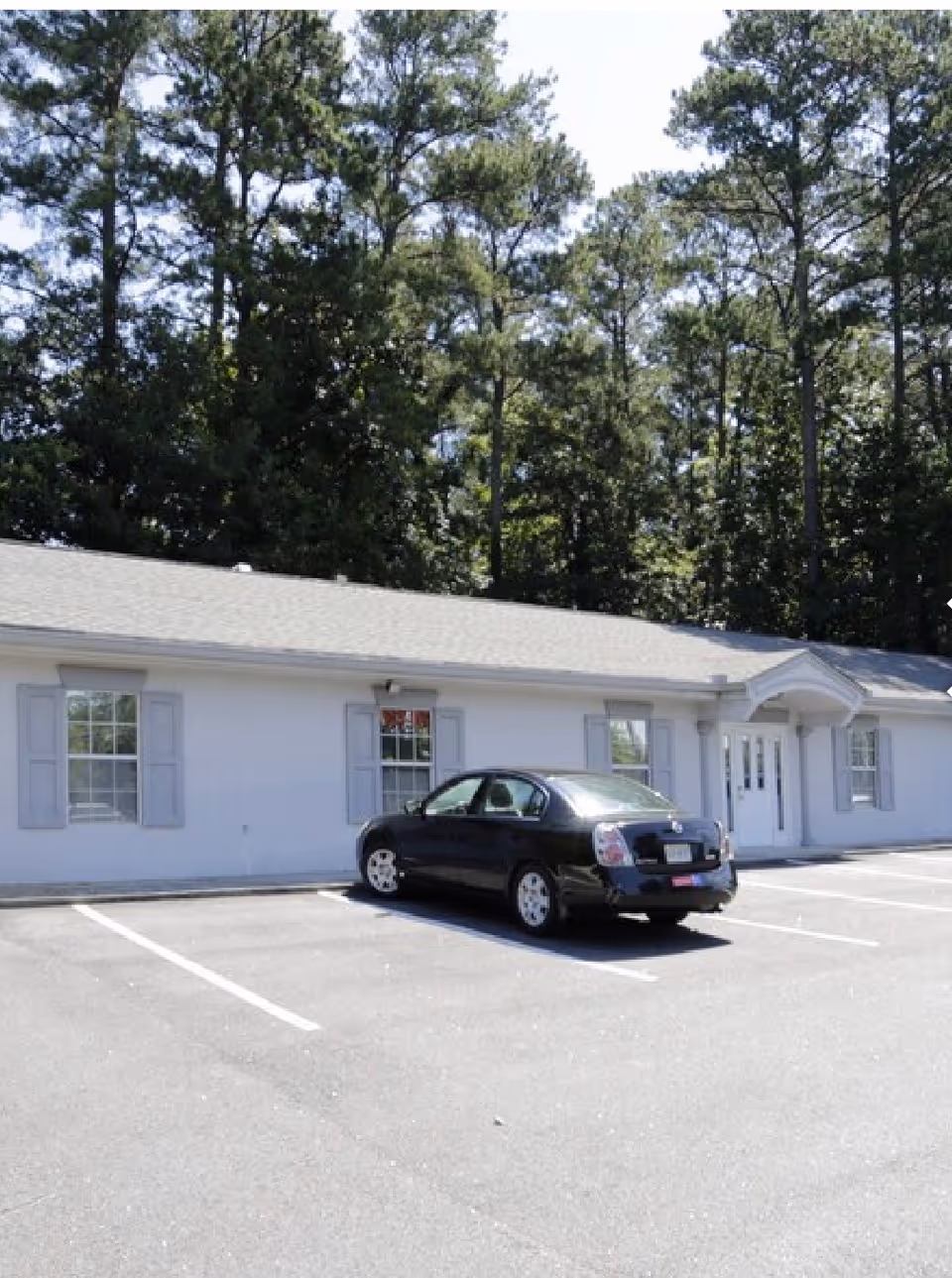 White single-story care facility front with blue shutters and a parked black car