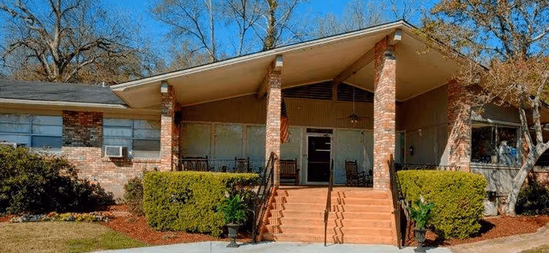 A brick senior facility with a covered front porch, steps, and manicured shrubs.