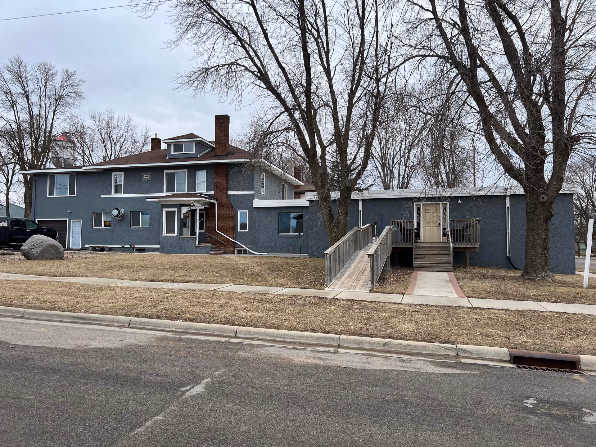 Blue two-story senior living building with ramp, stairs, and leafless trees along a street.