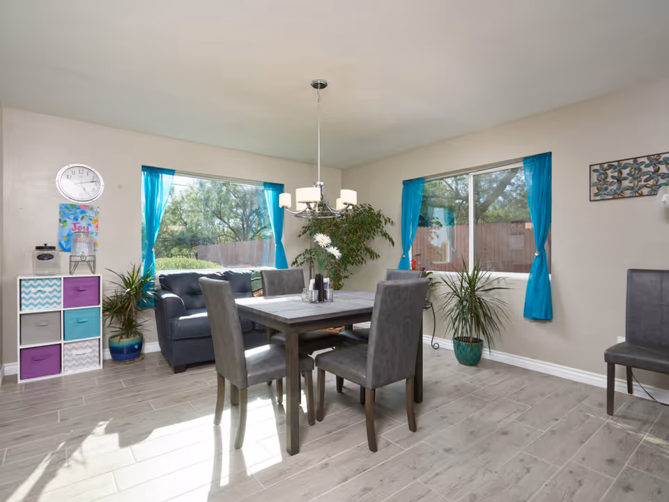 Bright combined dining and living area with a wooden table and gray chairs, a navy sofa, turquoise curtains, and potted plants by large windows.