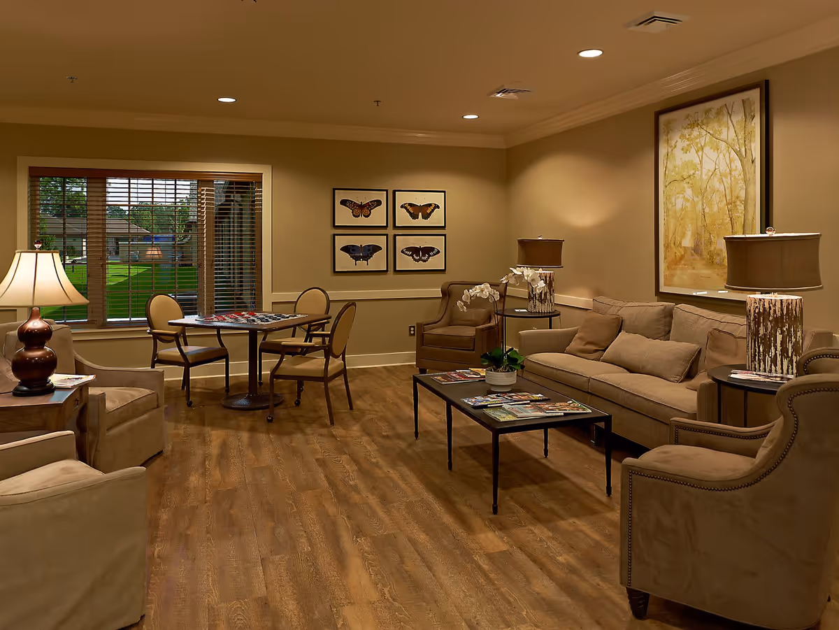 A cozy living room with beige walls and wooden flooring featuring a beige sofa, two armchairs, a coffee table with magazines and a potted plant, two side tables with lamps, a small round table with four chairs near a window with wooden blinds, and framed butterfly and nature artwork on the walls.