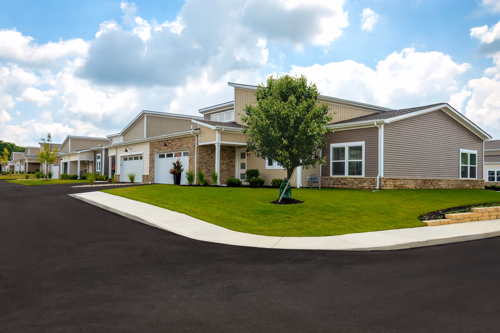 Exterior view of single-story residential buildings with garages, green lawns, and a tree along a paved road.