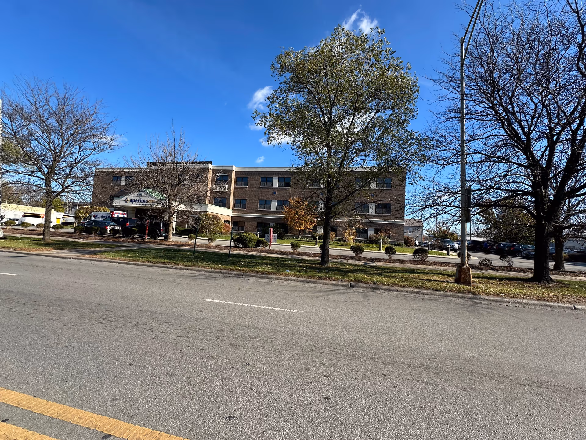 Exterior view of a three-story brick building with a sign reading Aperion Care International above the entrance. There are several trees with some leaves fallen, a parking lot with cars, and a clear blue sky with a few clouds.