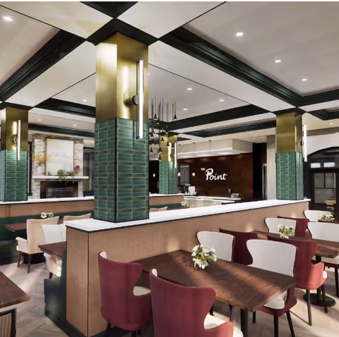 Interior view of a dining area at Brookwood Point featuring wooden tables with floral centerpieces, upholstered chairs in burgundy and beige, green tiled columns with brass accents, modern pendant lighting, and a fireplace in the background.