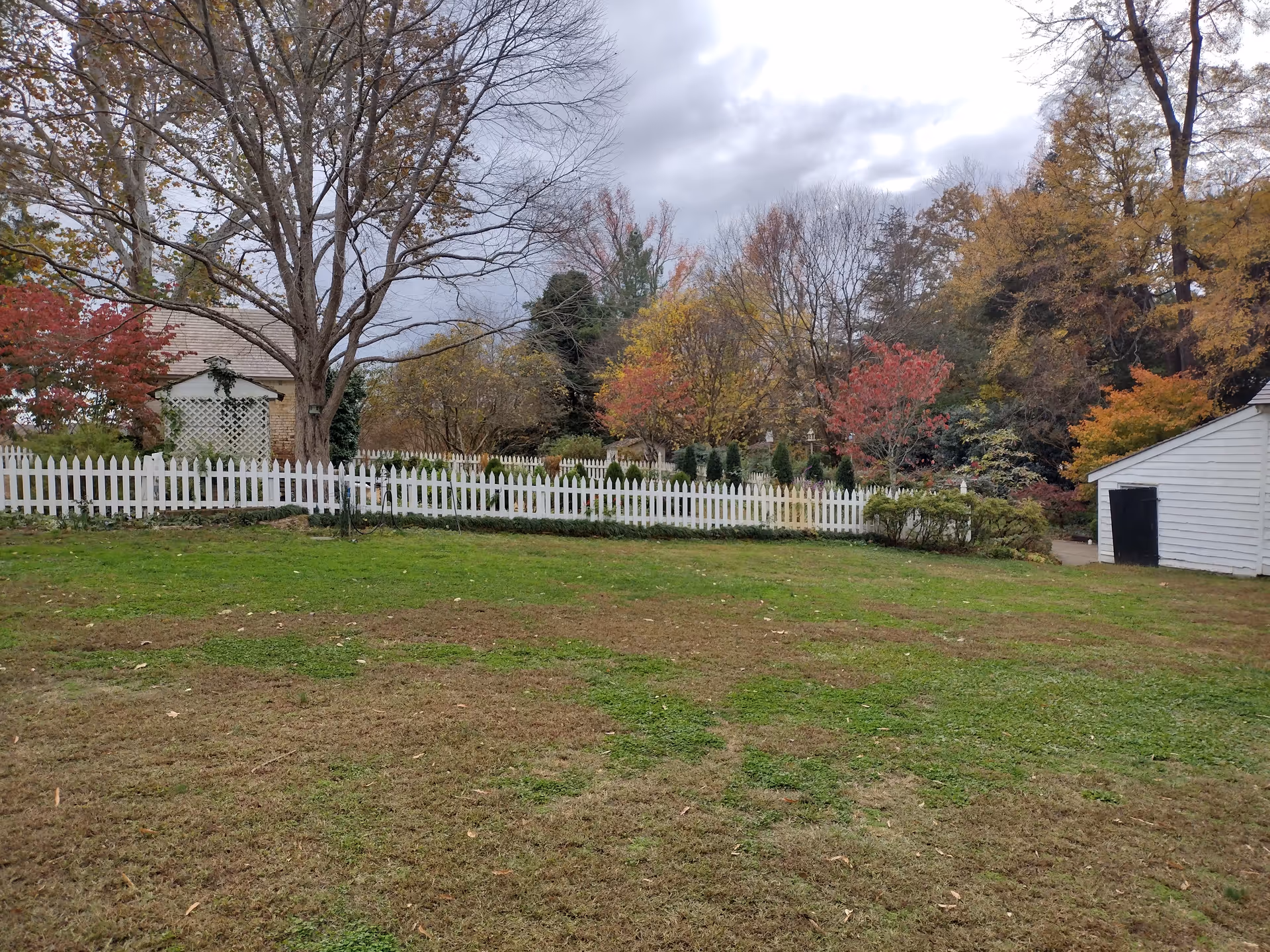 Grassy yard with a white picket fence, colorful autumn trees, and a small white shed.