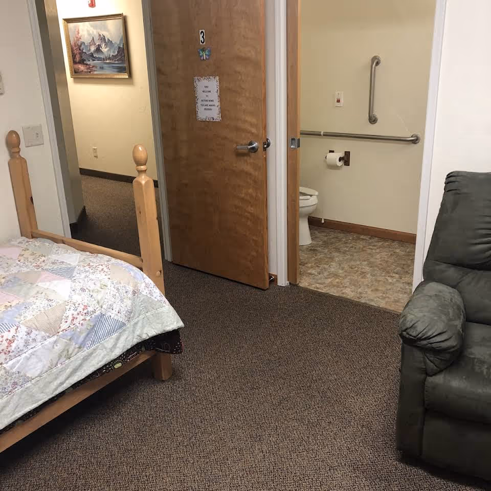 Interior view of a senior living facility room showing a wooden bed with a patchwork quilt, a green cushioned armchair, an open door leading to a bathroom with grab bars and a toilet, and another open door leading to a hallway with a framed painting on the wall.