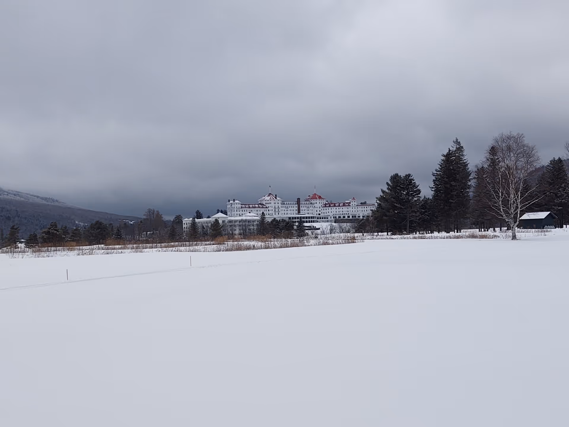 Snow-covered field with a distant large white building and trees under gray clouds