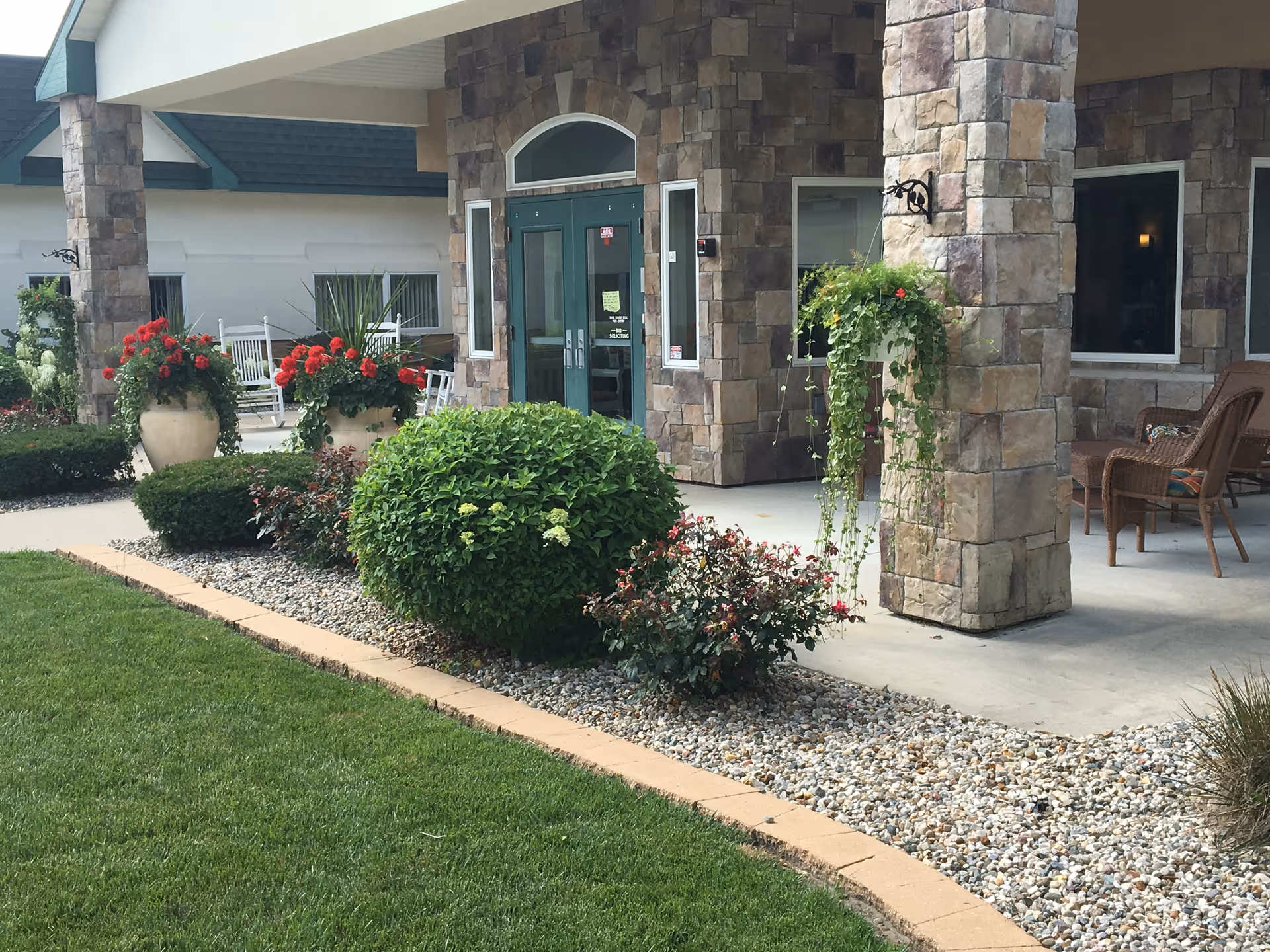 Covered front entrance with stone pillars, potted red flowers, hanging plants, wicker chairs and a landscaped lawn.