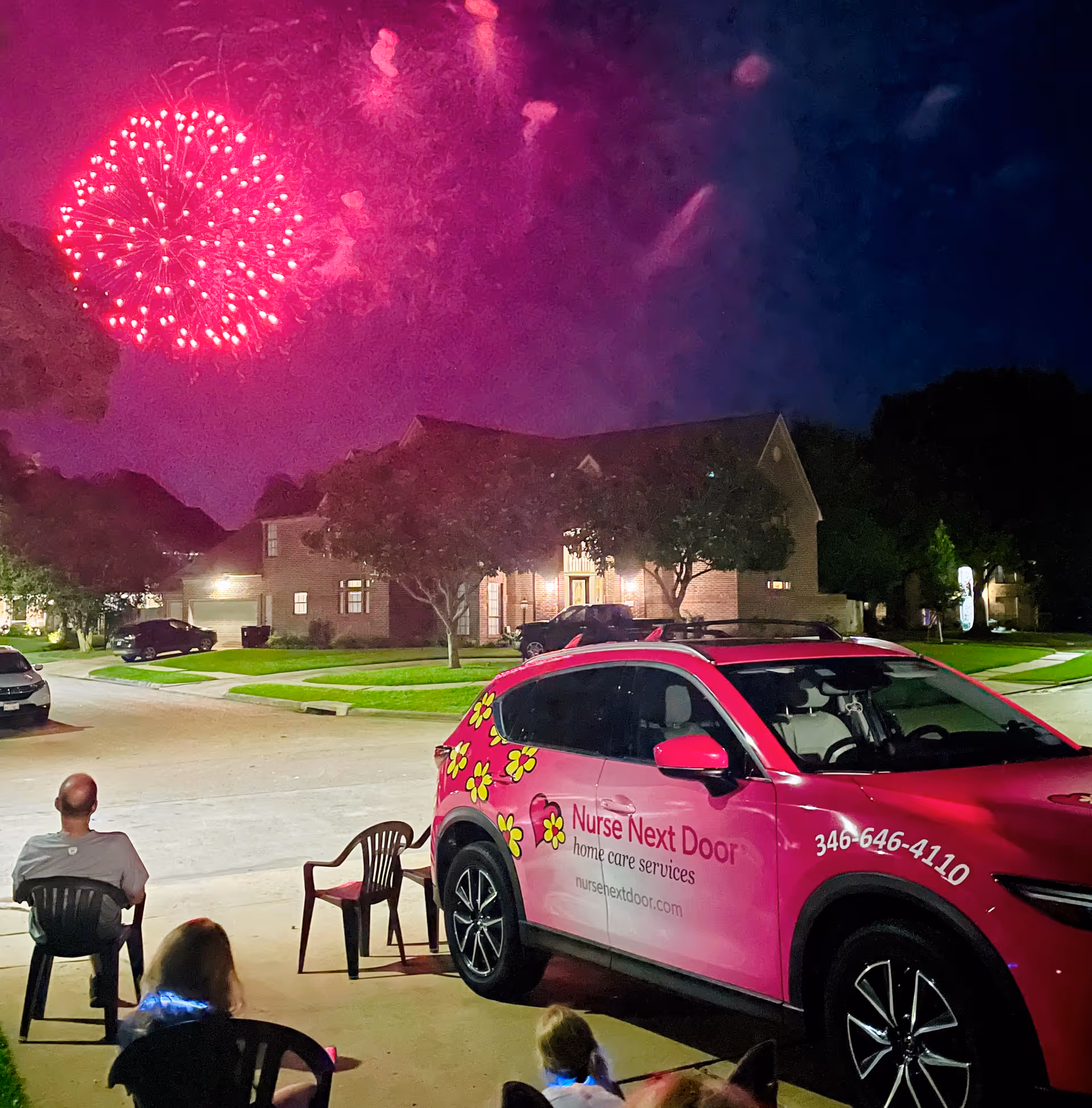 Pink Nurse Next Door van parked outside a home while residents sit in chairs watching fireworks.