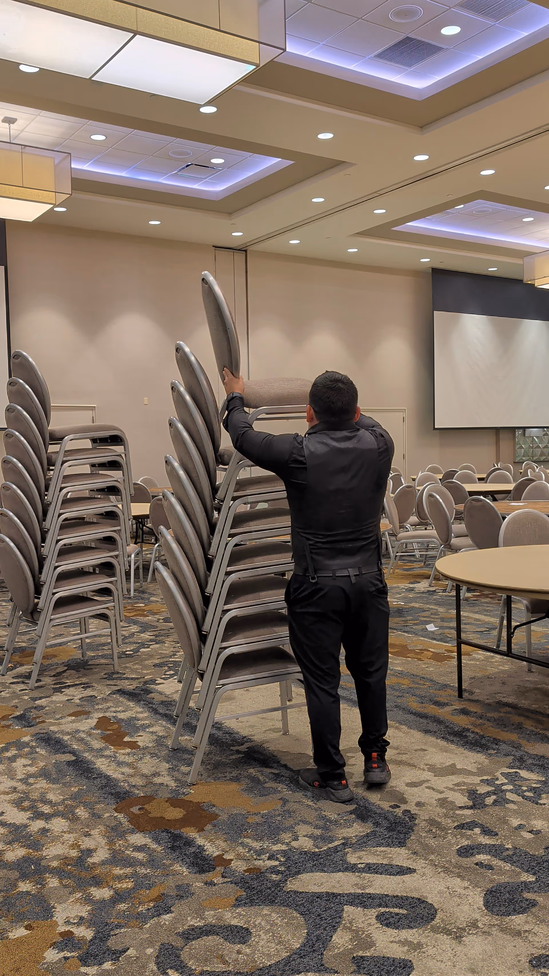 A staff member stacks gray stackable chairs in a spacious conference room.